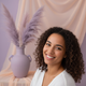 A happy young woman smiling warmly, looking directly at the camera, with a soft, blurred background in pastel colors.